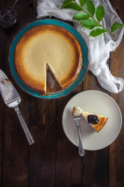 A slice of dense, pale New York-style cheesecake with a graham cracker crust on a white plate, the whole cheesecake visible in the background on a cake stand.