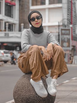 A stylish woman in a hijab poses in an urban setting, wearing sunglasses and casual attire.