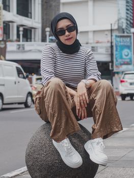 Stylish young woman in hijab sitting on street with urban fashion and trendy sunglasses.