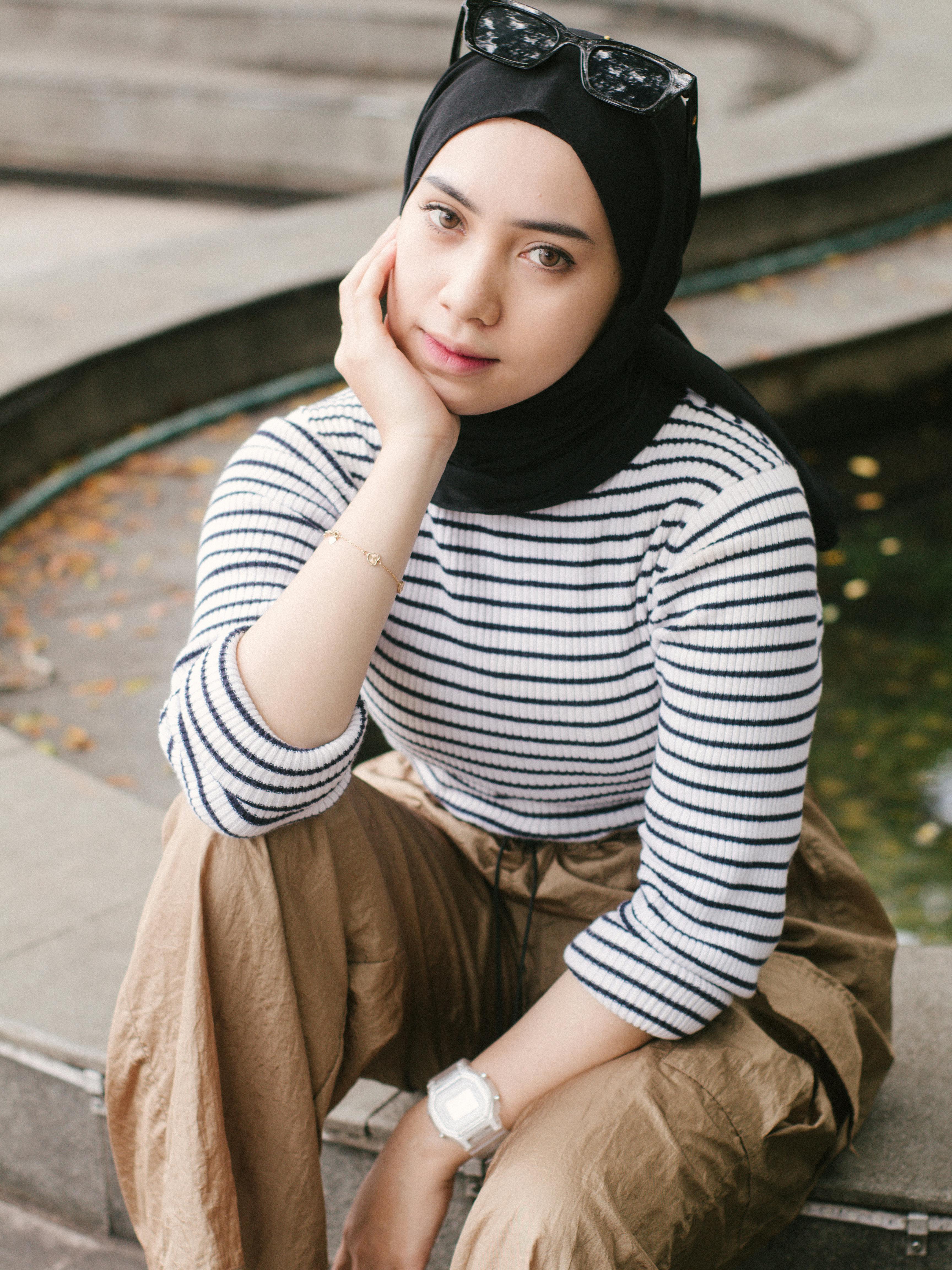 Fashionable young woman wearing a headscarf, sitting by a fountain in an urban setting.