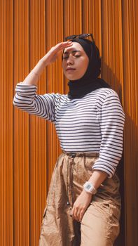 Woman in striped fashion leaning on copper wall, shielding face from sunlight in vertical shot.