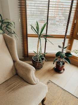 A cozy modern interior featuring a beige armchair, potted plants, and large windows.