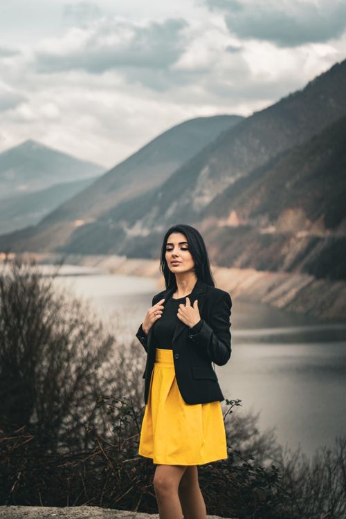 Free Woman poses by scenic lake and mountains, wearing a yellow skirt and black jacket. Stock Photo