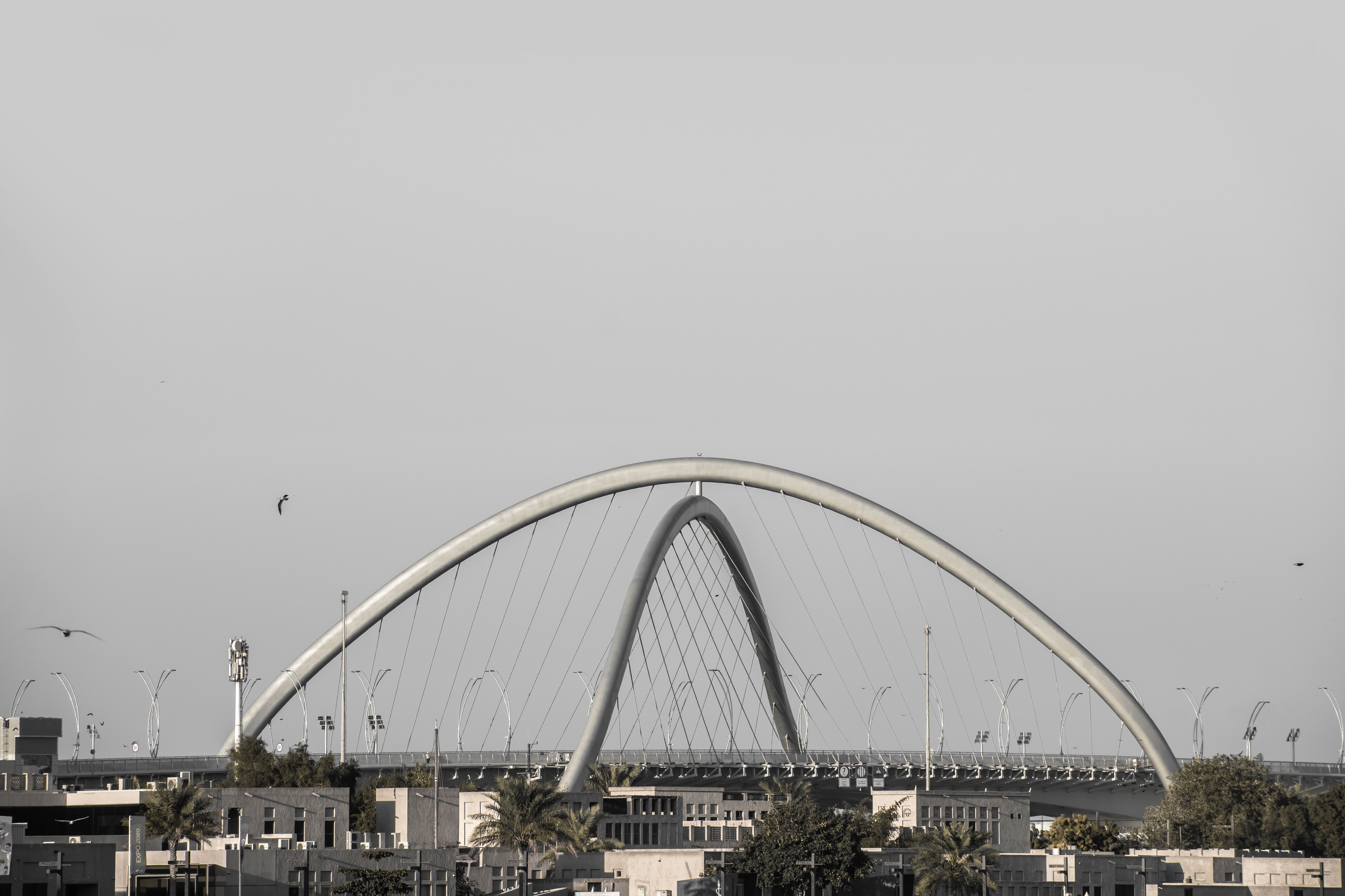 A modern arch bridge spans over Dubai's urban landscape with a clear sky background.