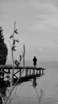 Silhouette of a person standing on a pier over a calm lake under an overcast sky.