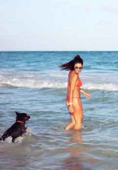 A woman in a red bikini and sunglasses enjoys a sunny beach day with her dog in the ocean.