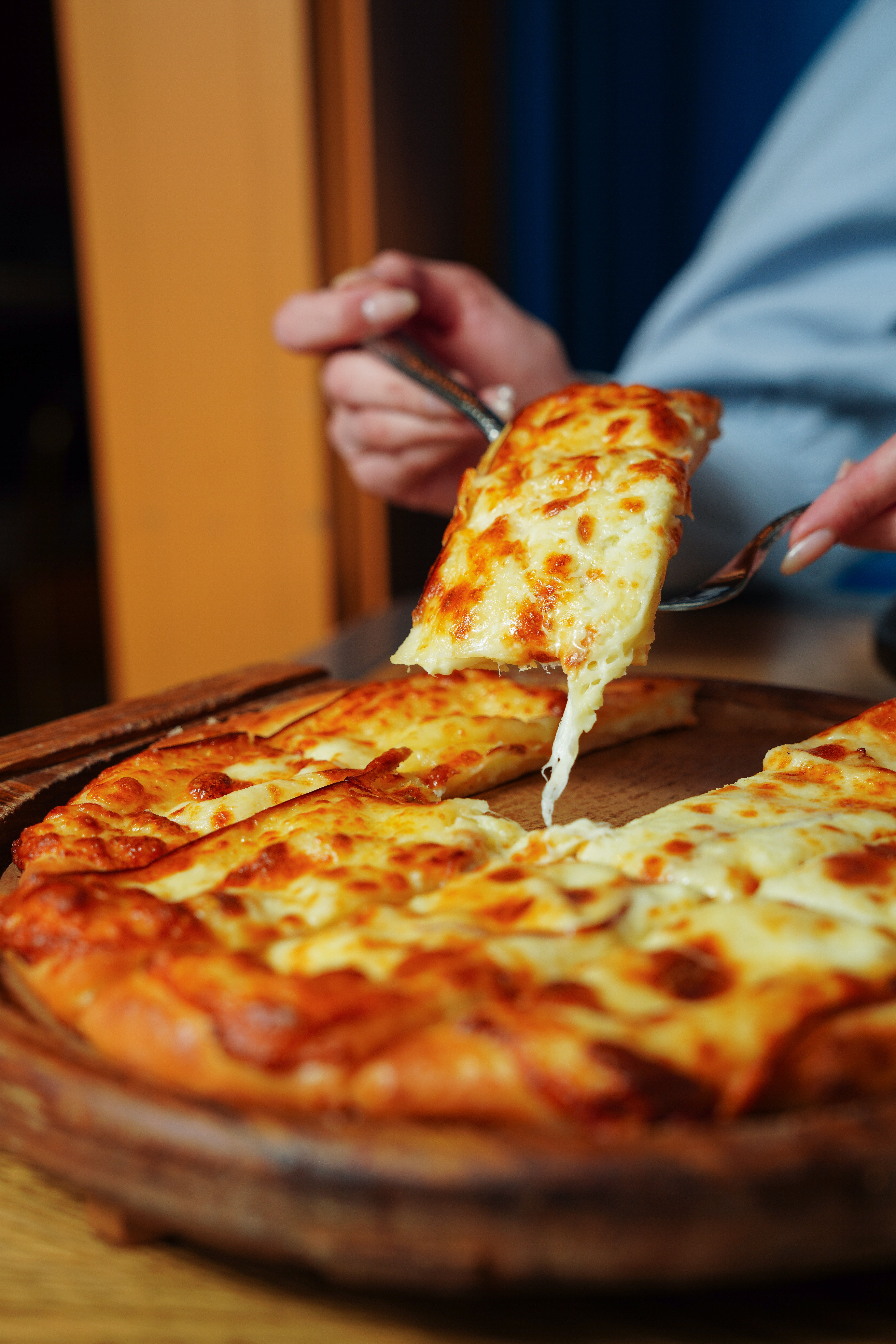 Close-up of a hot cheese pizza slice being lifted from a wooden platter indoors.
