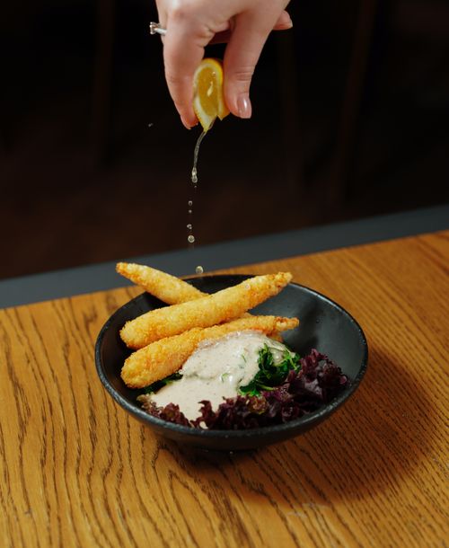 Free A vertical shot of crispy fish sticks with lemon drizzle, served with sauce and greens in a black bowl. Stock Photo