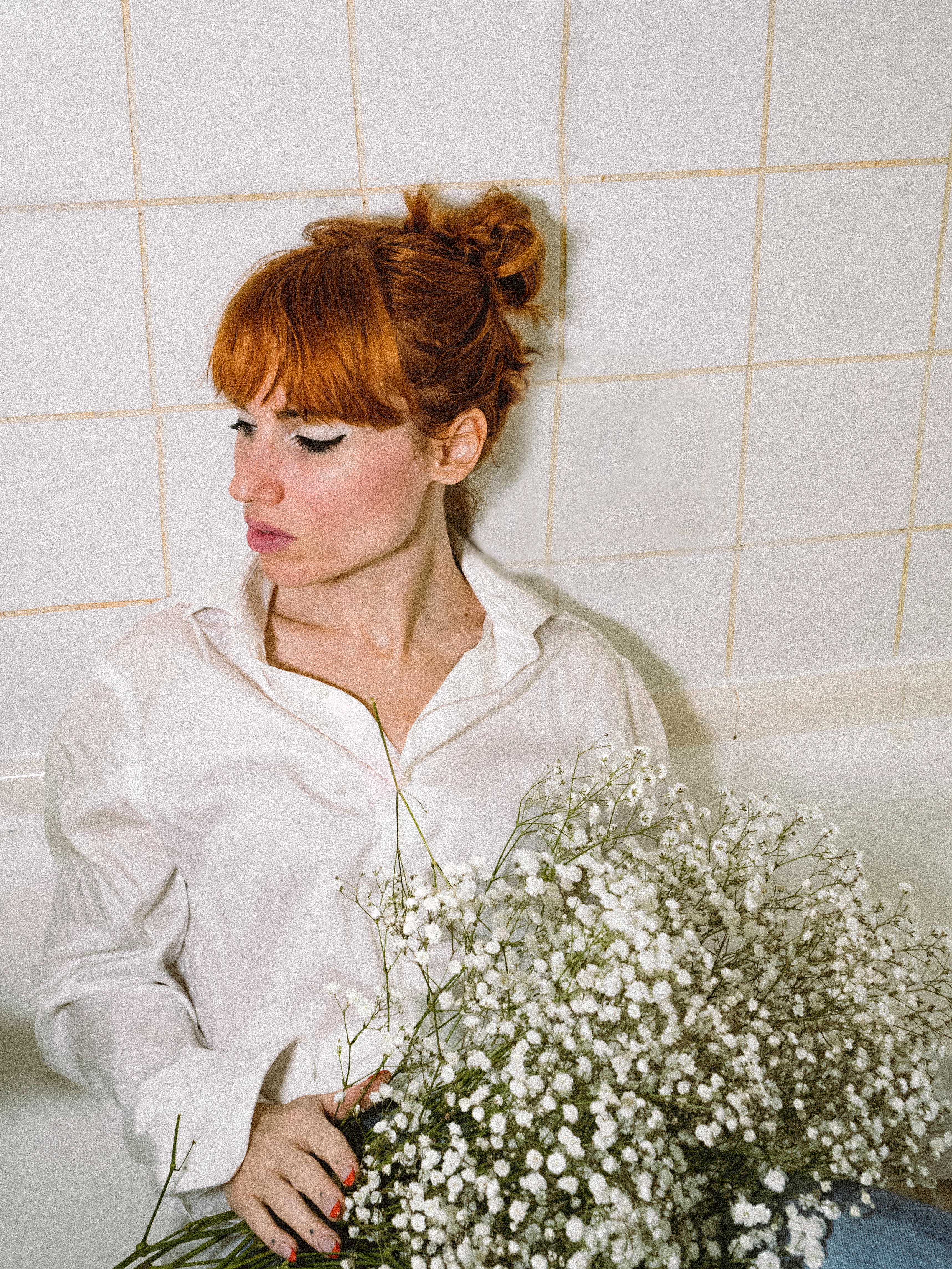 A redhead woman in white shirt poses with flowers against a tiled background, embodying elegance and tranquility.