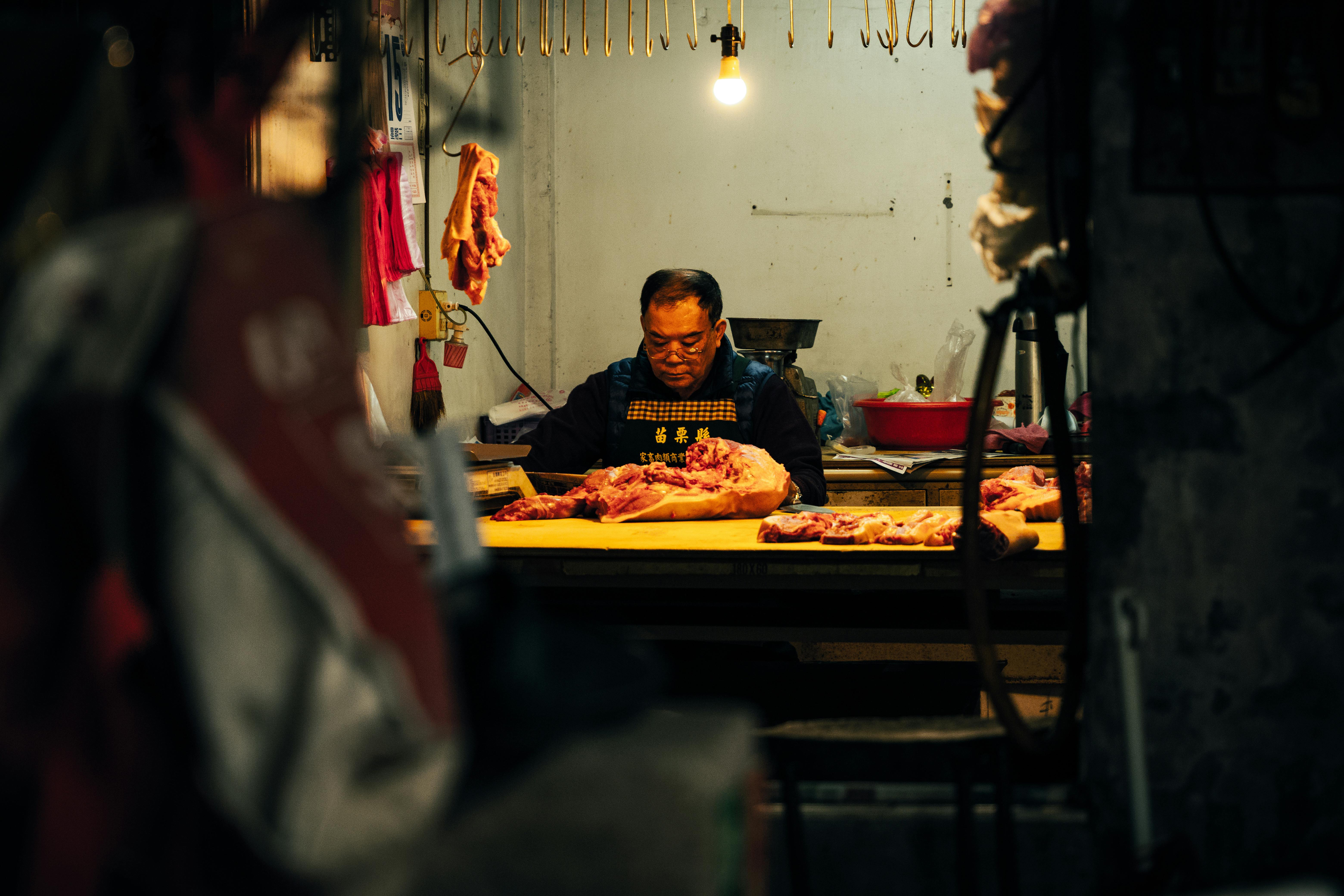 A dedicated butcher cuts meat in a dimly lit indoor setting, focused on his work.