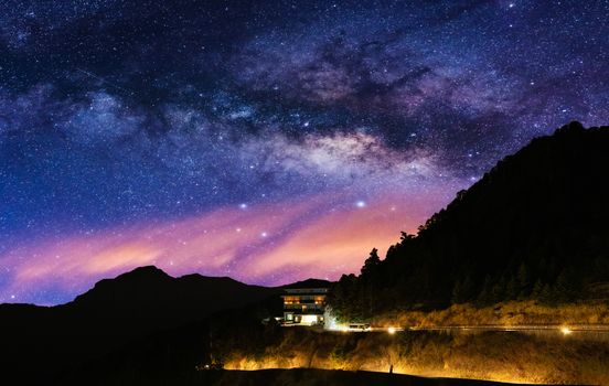Stunning view of the Milky Way over a serene mountain landscape with a lit house at night.