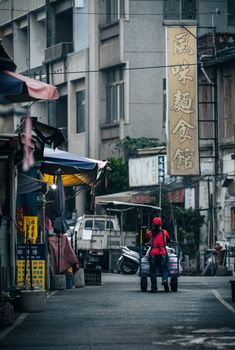 A street vendor pushes a cart through a bustling Asian urban market, surrounded by buildings and signs.