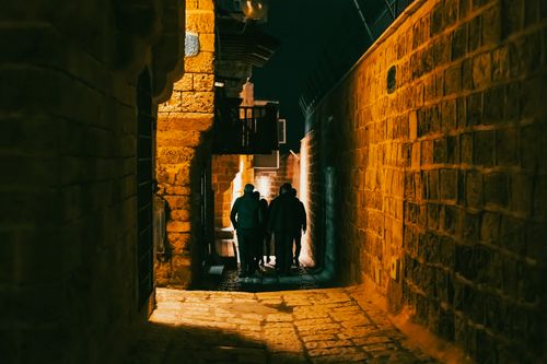 Free Silhouetted figures walk through the warmly lit alley of Tel Aviv-Jaffa at night. Stock Photo