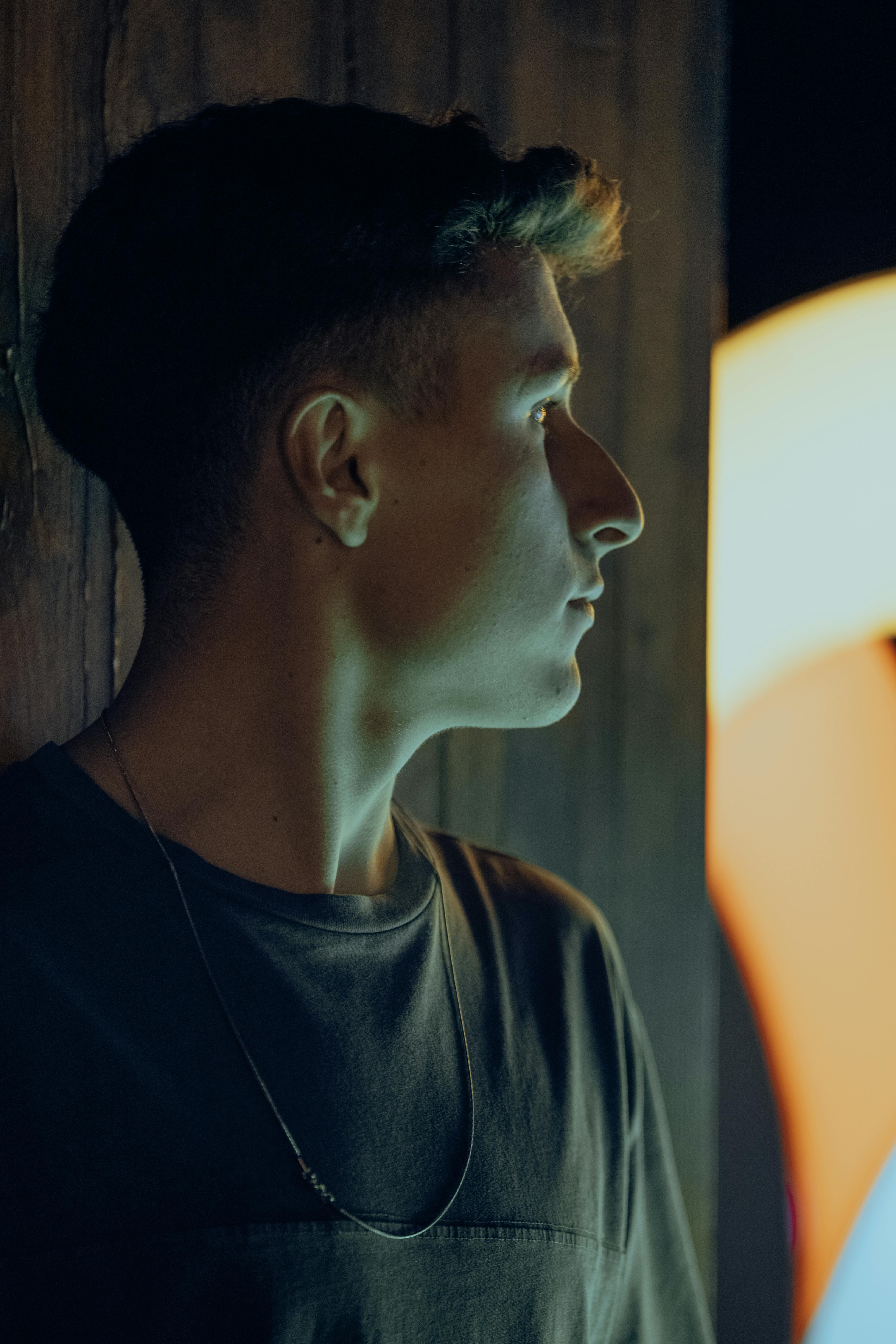 Side profile of a man with short hair and necklace in dim lighting, evoking a moody atmosphere.