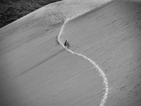 Aerial view of two people walking on a large sand dune, casting shadows in the landscape.