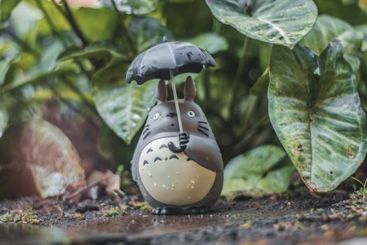 A toy figure holds an umbrella amidst raindrops and lush green leaves in a garden setting.