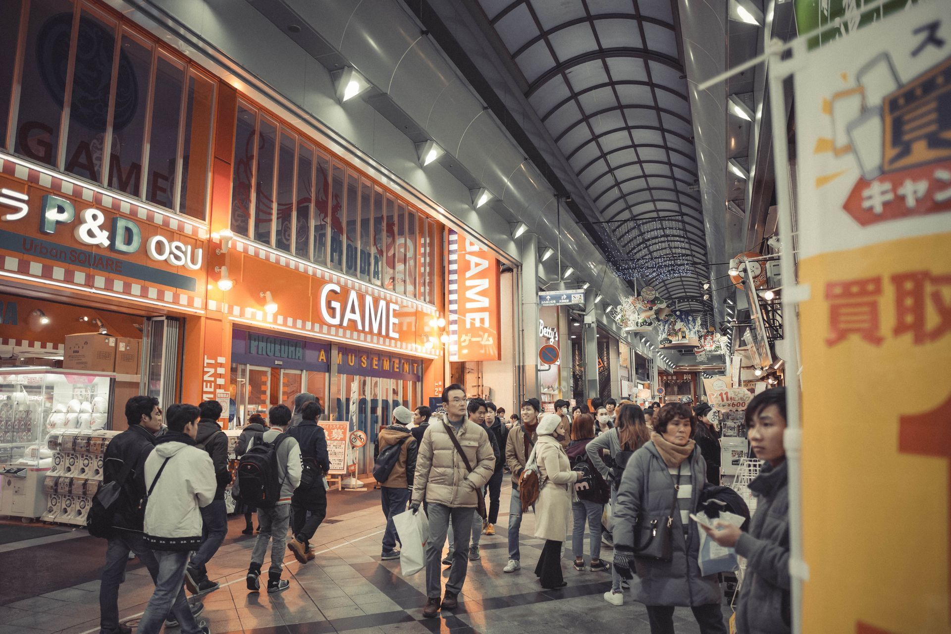 Crowded shopping arcade scene with people exploring shops under bright lights.
