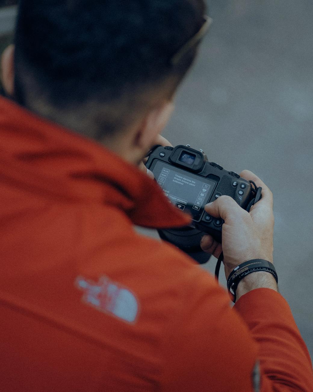 A man in a red jacket reviews camera settings while holding a DSLR outdoors.