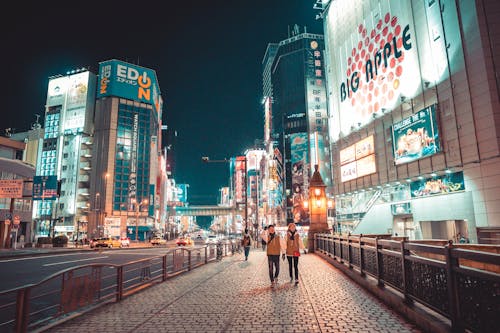 Free Couple Holding Hands While Walking on Pathway in Front of Big Apple Building Stock Photo