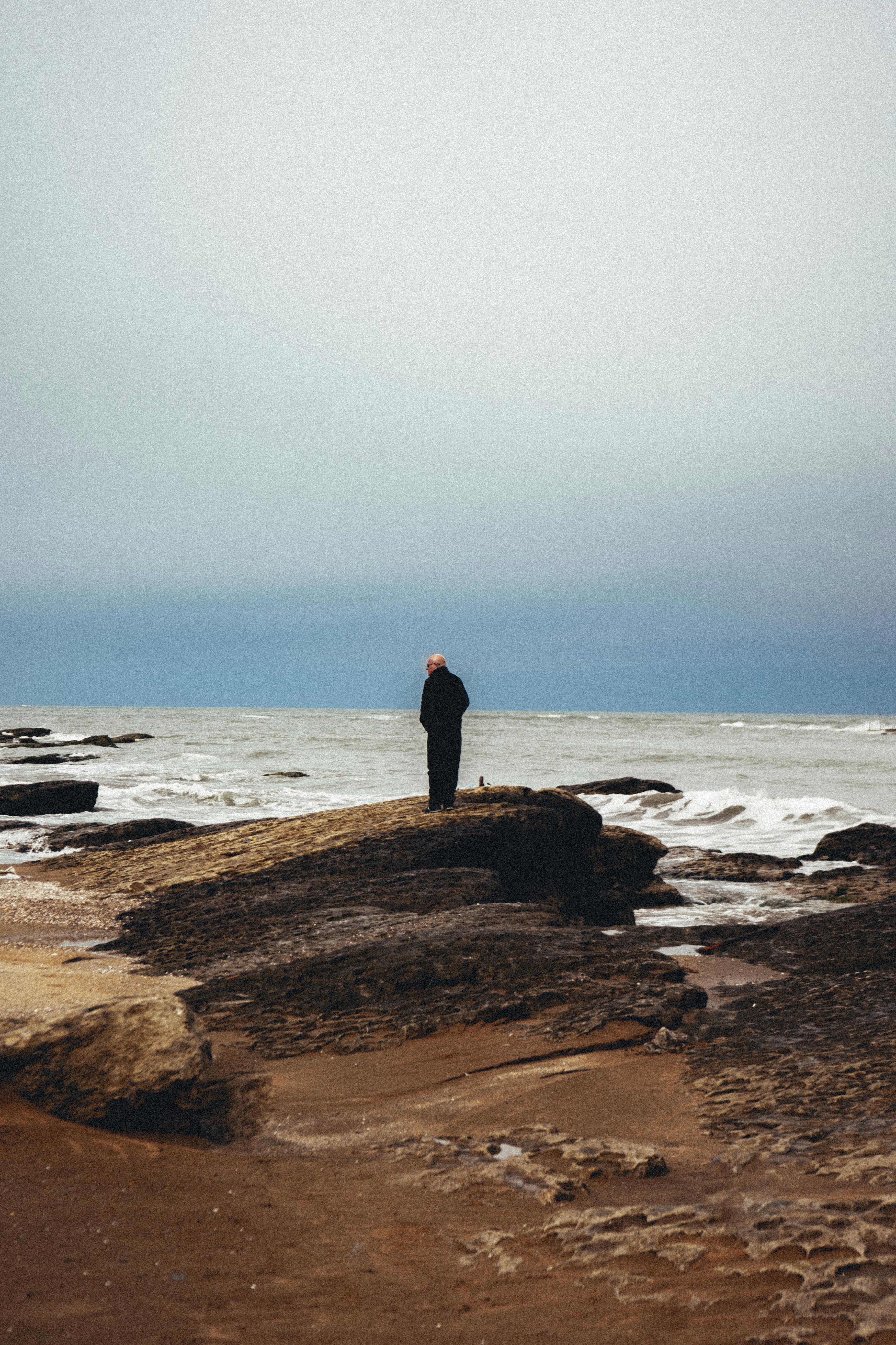 Elderly man standing alone on a rocky Baku coastline under an overcast sky.