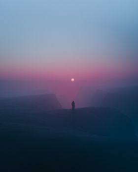 A lone figure stands against a misty cliffside with the moon visible at twilight.