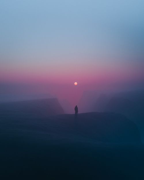 Free A lone figure stands against a misty cliffside with the moon visible at twilight. Stock Photo