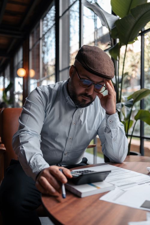 Free Businessman in a modern office setting using a calculator, surrounded by plants and documents. Stock Photo