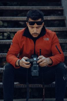 A man in a red jacket sits on stairs, focused on his camera outdoors.