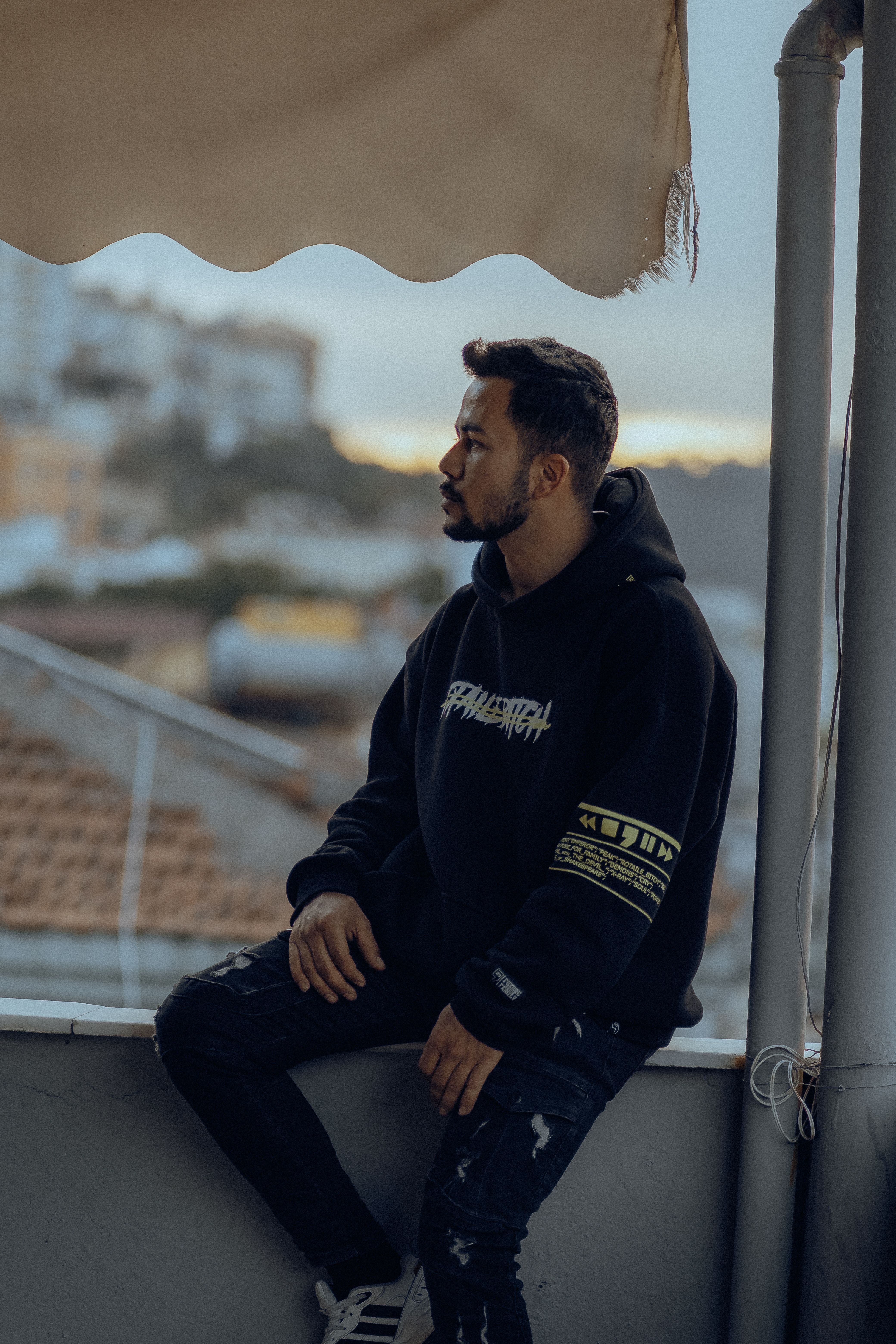 Portrait of a young man in a hoodie sitting on a rooftop in the city during twilight.