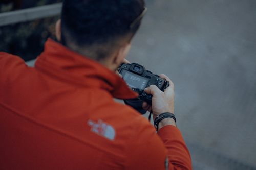 Free Back view of a man in a red jacket adjusting settings on a DSLR camera. Stock Photo