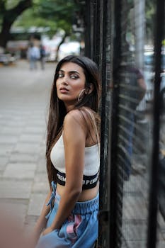 A fashionable woman poses by a fence on a city street, exuding confidence and style.