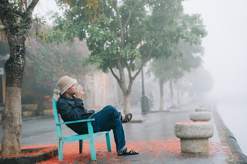 Man Sitting on Chair and Smoking near Trees in Town under Fog