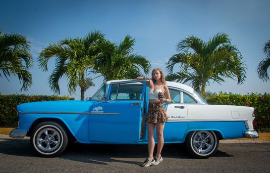 A young woman stands by a vibrant blue Chevrolet Bel Air with palm trees in the background.