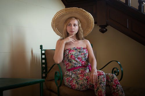 Young Woman in a Dress with a Floral Pattern and a Hat Sitting on a Chair in a Vintage Interior 