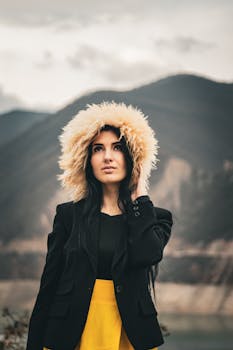 Fashionable woman in fluffy hat and elegant outfit posing outdoors with mountains in the background.