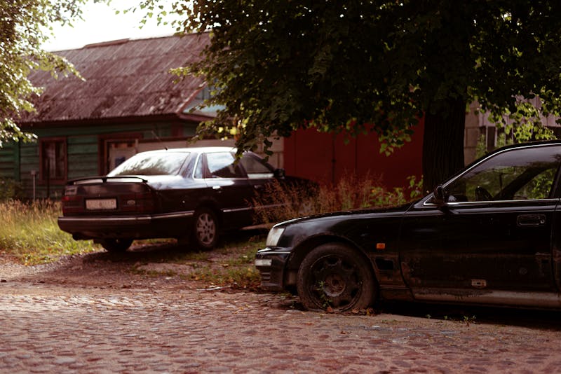 Two black cars in a neglected suburban driveway beside an old house.