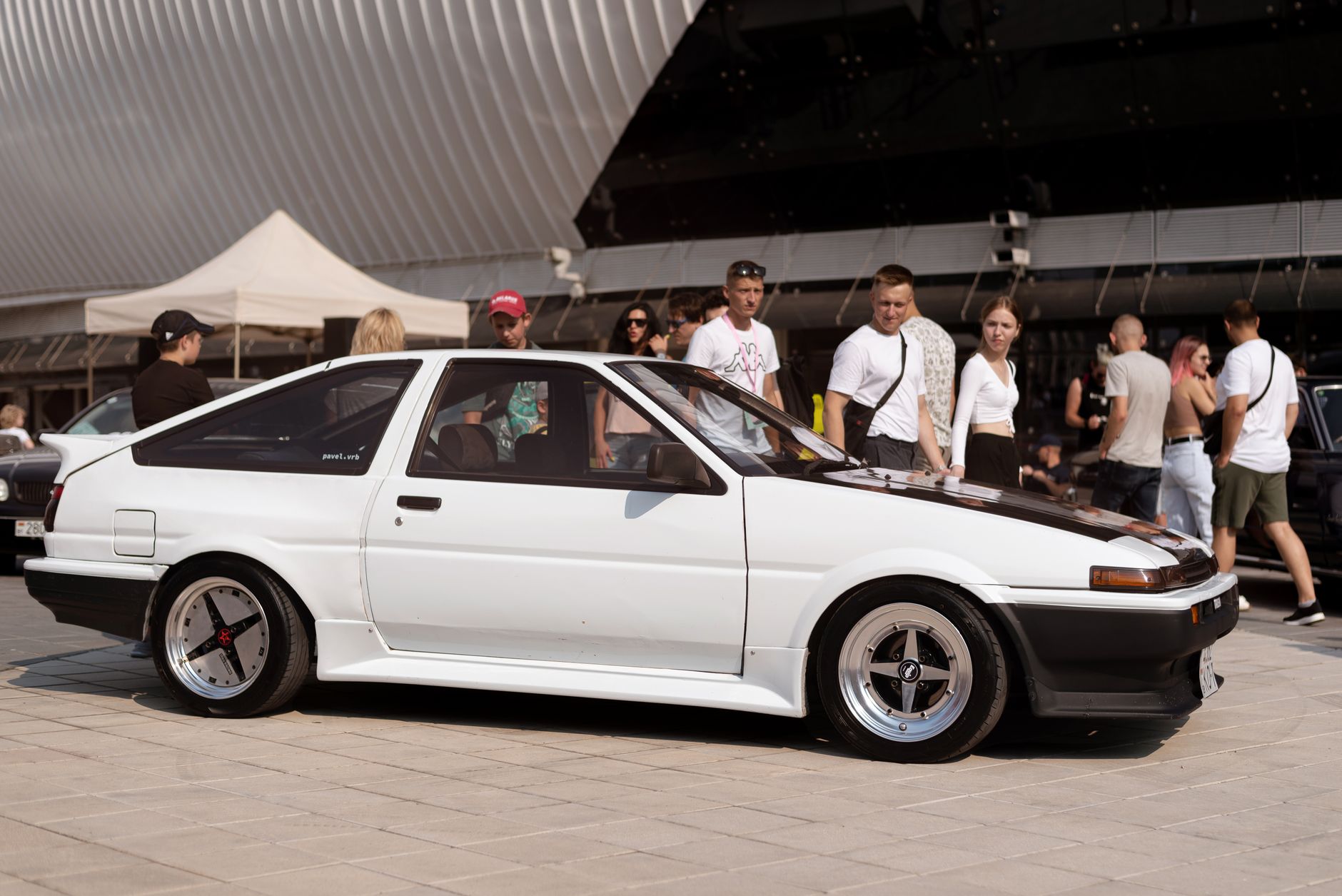A vintage white car on display at an outdoor auto exhibition surrounded by people.