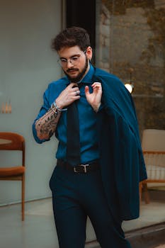 A fashionable young man with tattoos adjusts his tie while holding a jacket, showcasing modern style.