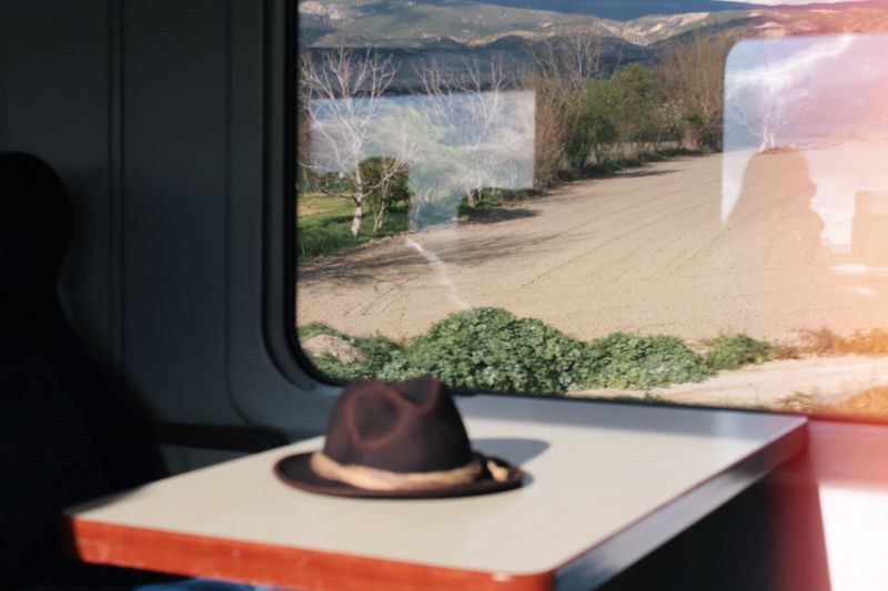 Inside a train carriage, a lone hat rests on a table with fields visible through the window.