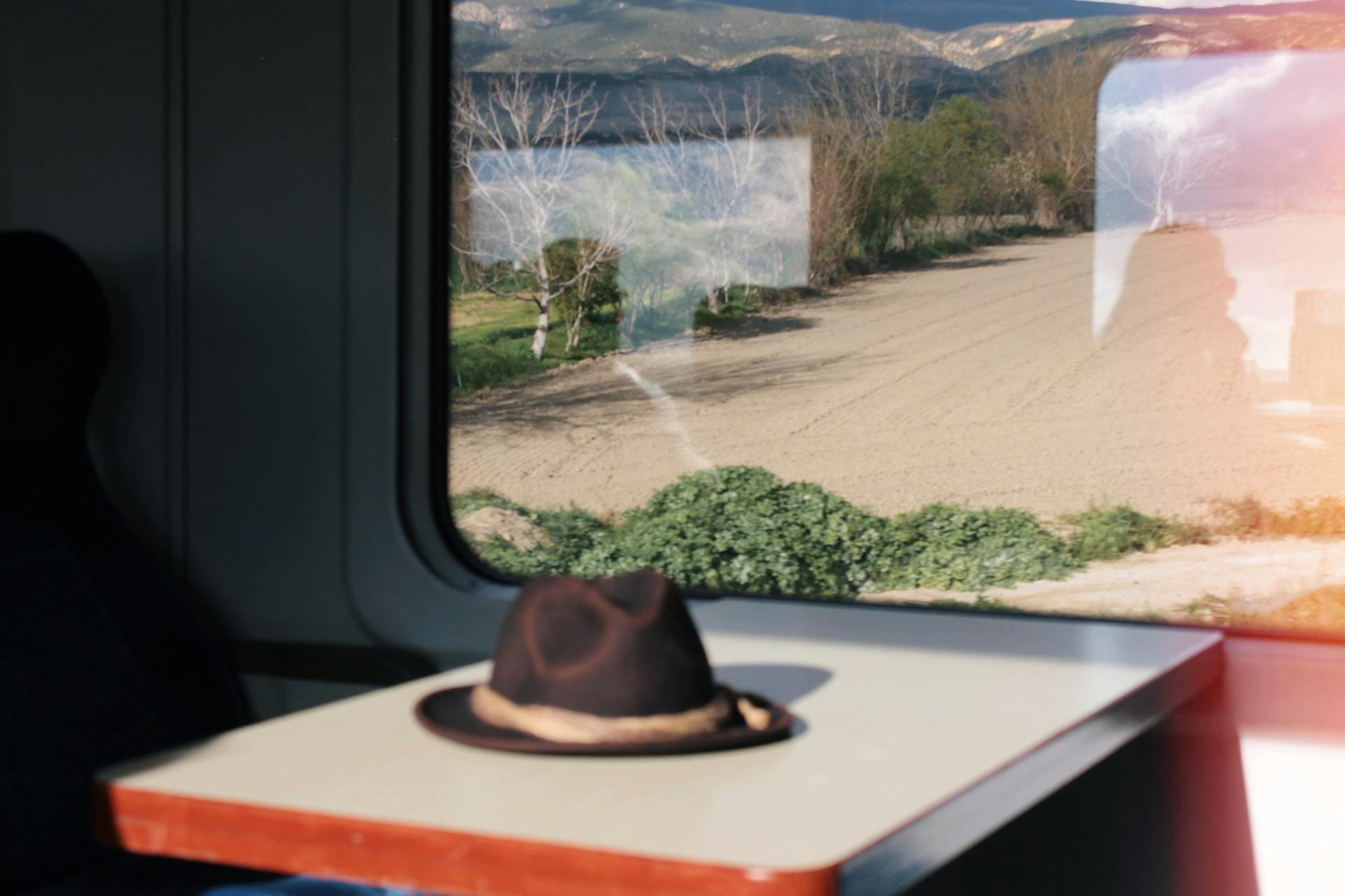 Inside a train carriage, a lone hat rests on a table with fields visible through the window.