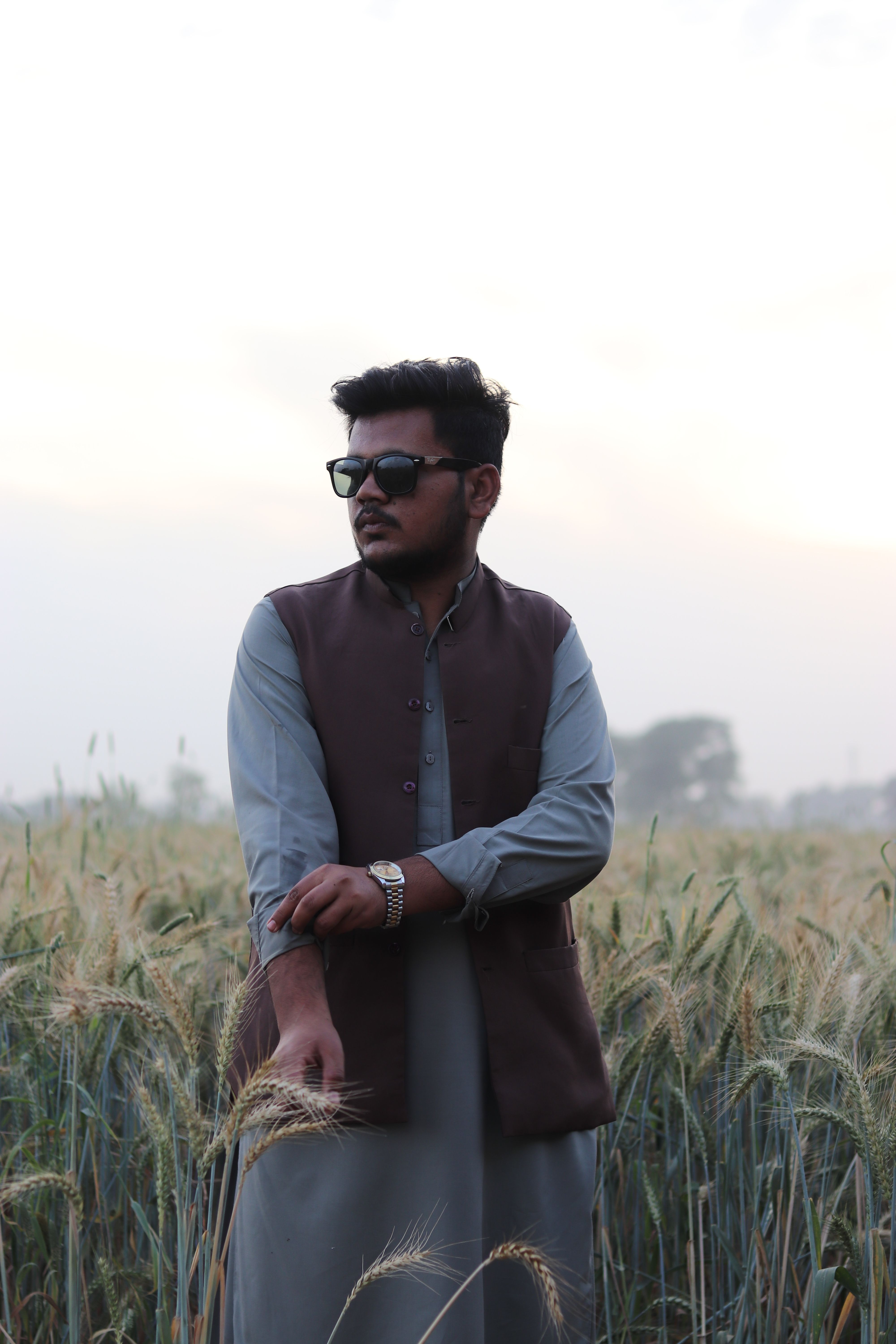 A fashionable man wearing sunglasses poses confidently in a rural wheat field during early evening light.