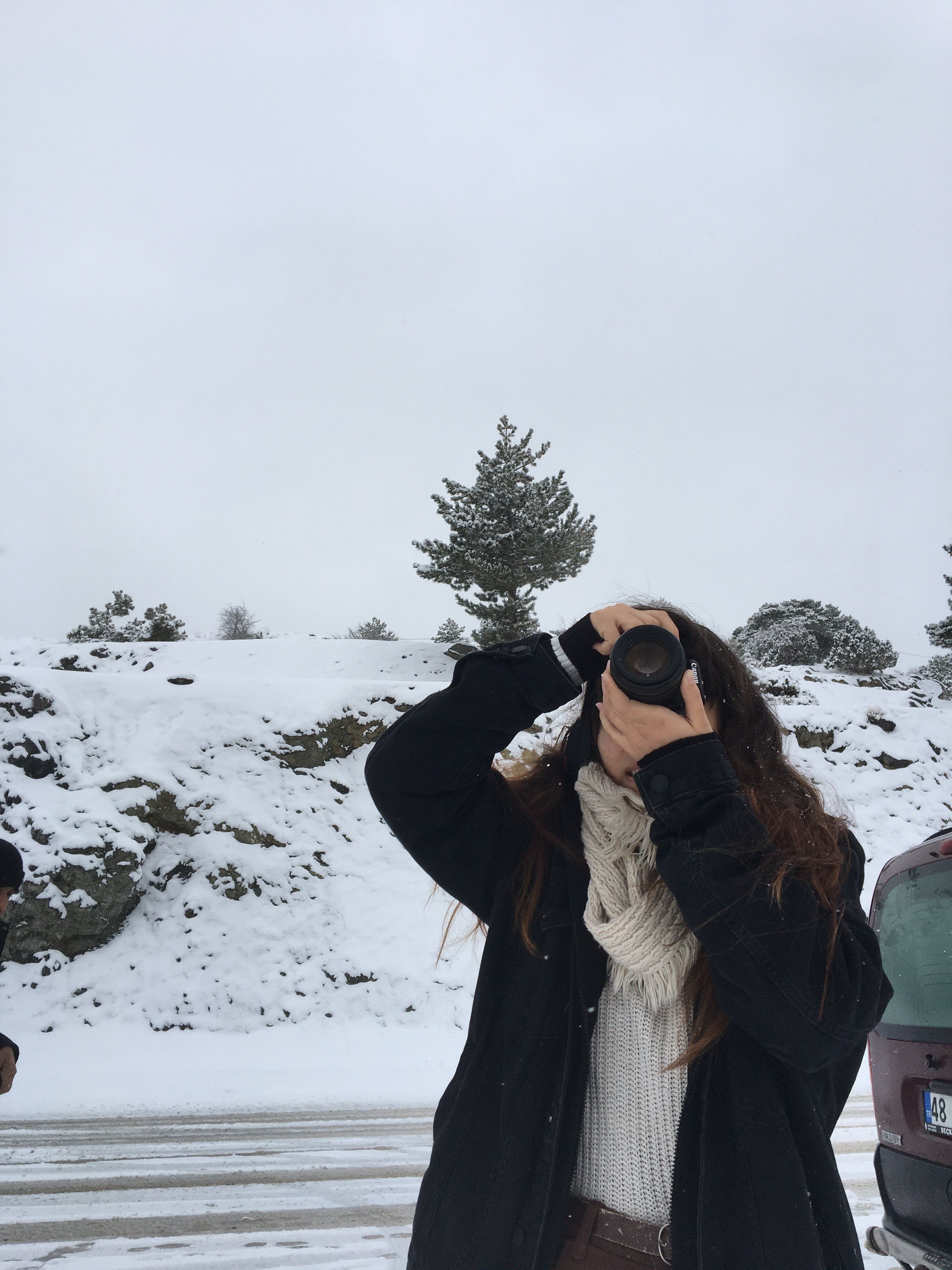 A woman photographs a snowy landscape wearing winter attire. Overcast skies set a serene mood.