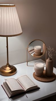 A cozy desk scene featuring a lamp, open book, and decorative elements like a mirror, candle, and flower vase.