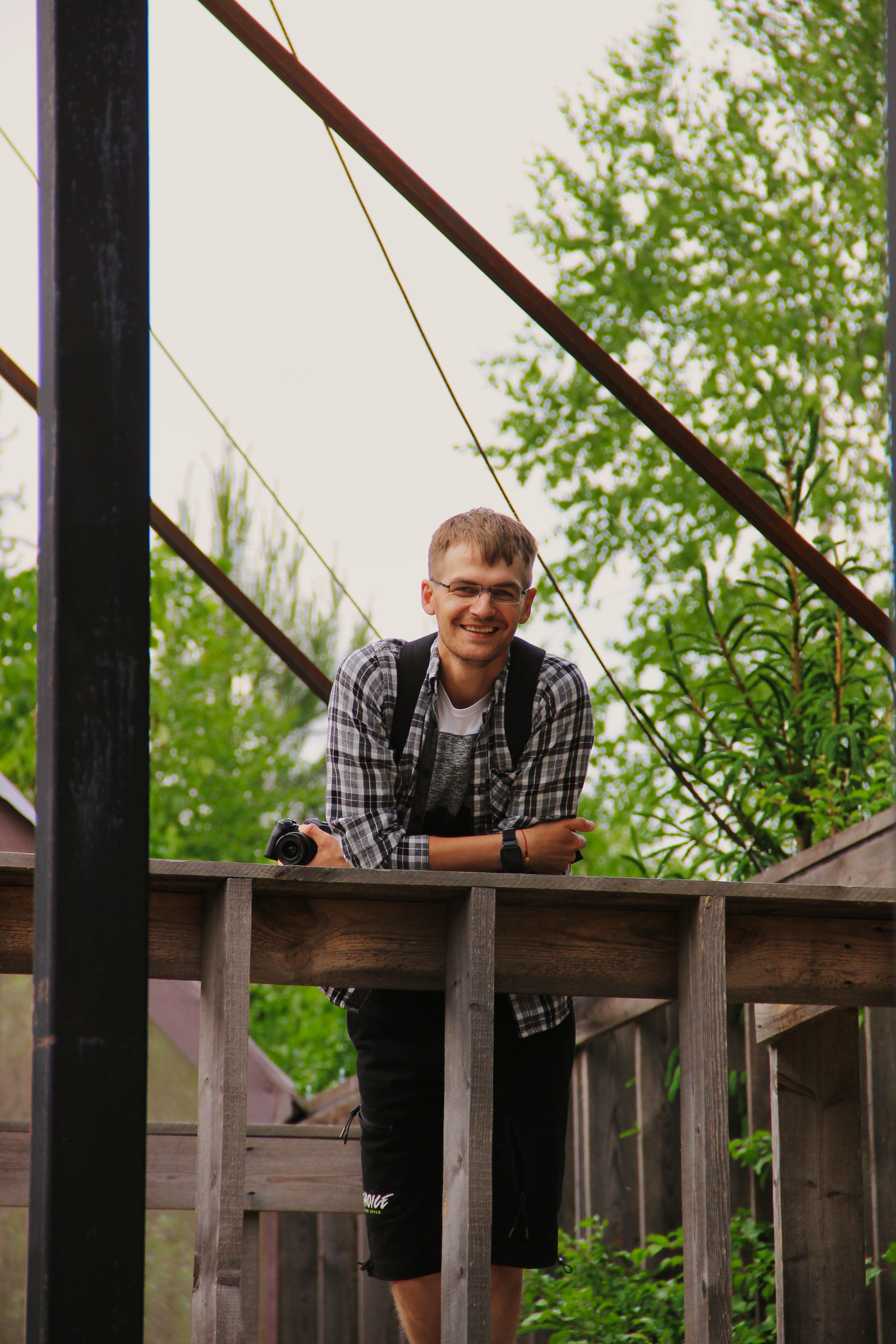 Smiling young man with camera leaning on a wooden railing outdoors, surrounded by greenery.