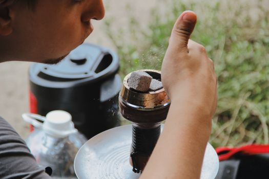 A man preparing hookah by blowing charcoal pieces to ignite them outdoors.