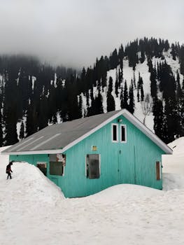 A tranquil scene of a snowy mountain cabin surrounded by pine trees under a cloudy sky.