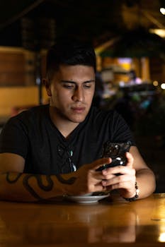 Young man engrossed in his smartphone at a cozy nighttime café, creating a relaxed ambiance.