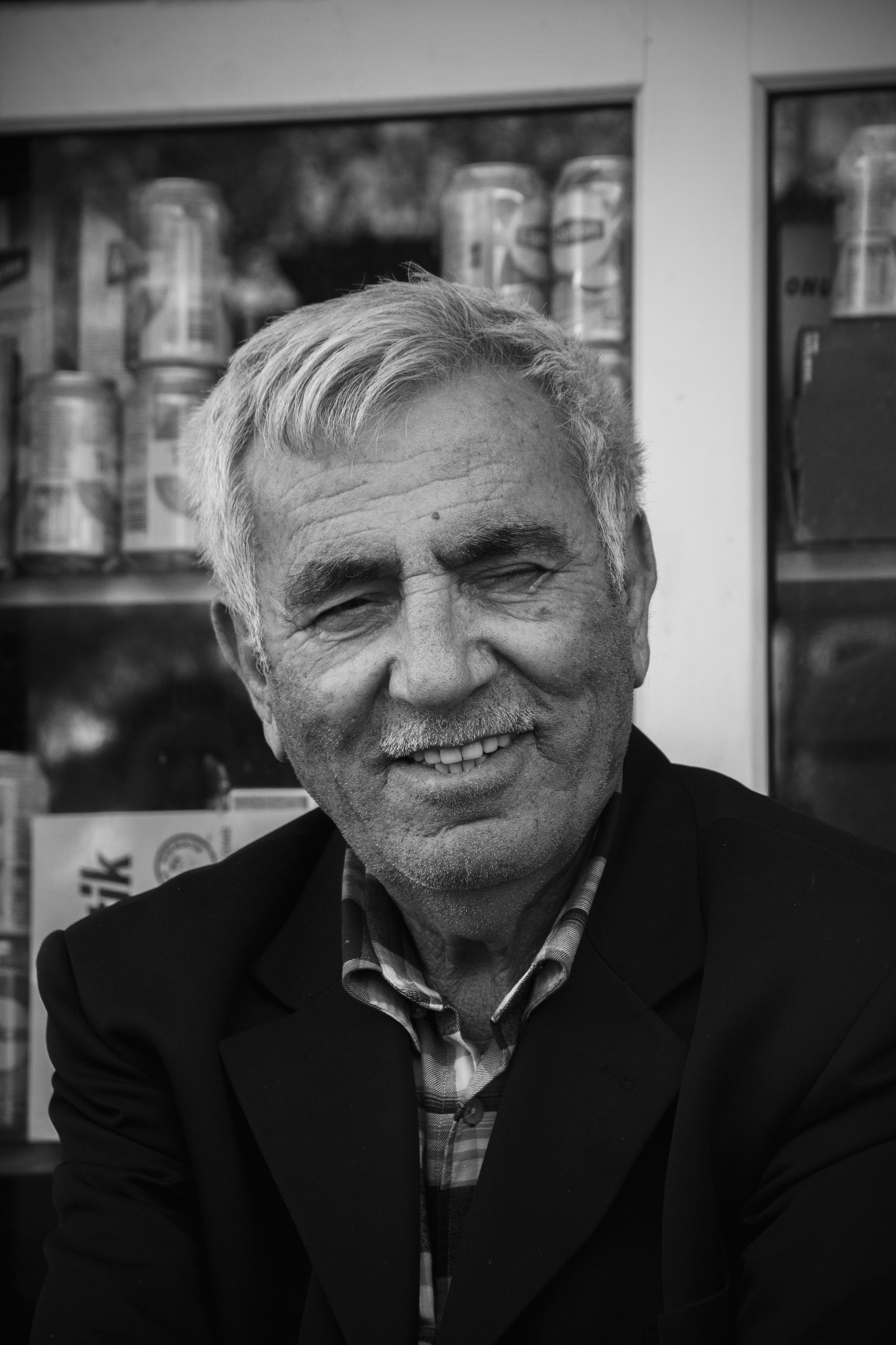 A senior adult man with a mustache smiles in a black and white portrait set in Adana, Türkiye.