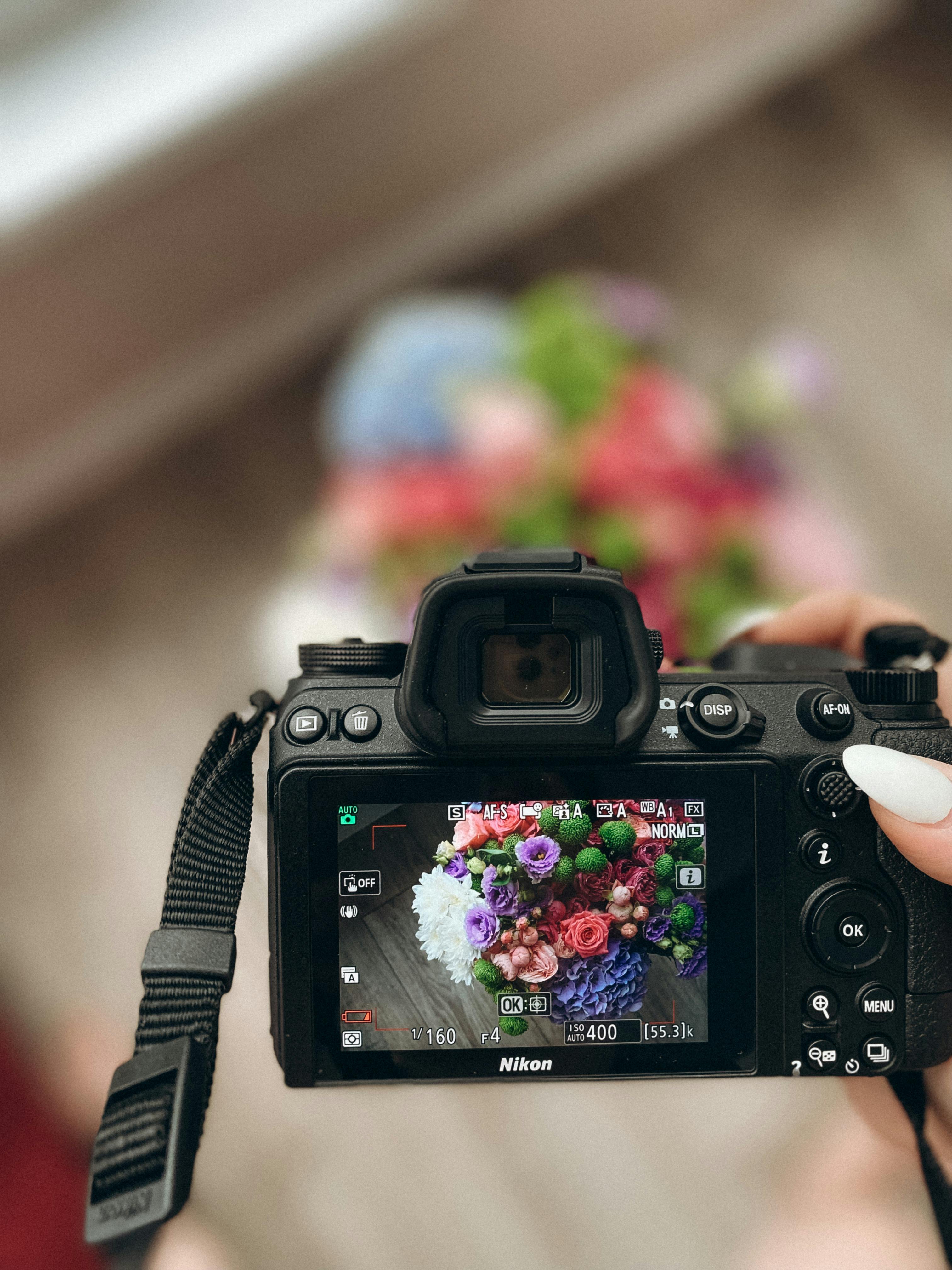 A hand holding a camera photographs a vibrant and colorful flower bouquet indoors.