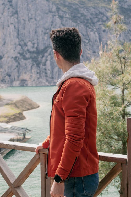 Free Man Wearing an Orange Jacket Standing on a Bridge Overlooking a Lake Stock Photo